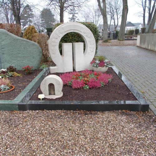 Grabstein aus beigem Kalkstein in Form des griechischen Buchstabens Omega. Die Rundung ist bis auf eine kleine Fläche für die Beschriftung mit Rosen verziert. Die Einfassung ist aus Impala.