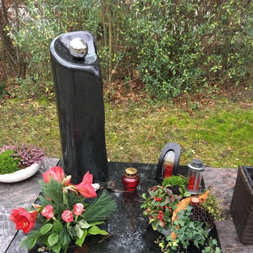 Eine Säule in geschwungener Form mit Abdeckund aus schwarzem Naturstein, in der eine Edelstahlkugel in der oberen Aushöhlung befestigt ist.