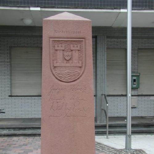 Ein Obelisk aus rotem Sandstein vor dem Rathaus in Niederkassel der die Partnerstädte zeigt.